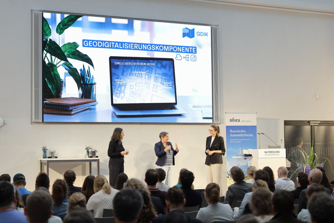Vorstellung der Geodigitalisierungskomponente Konferenzsaal mit drei Frauen auf der Bühne unter einer großen Leinwand, auf der unter dem Titel